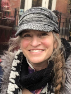 Woman with blonde hair in braids and a grey crap and black scarf looking at the camera